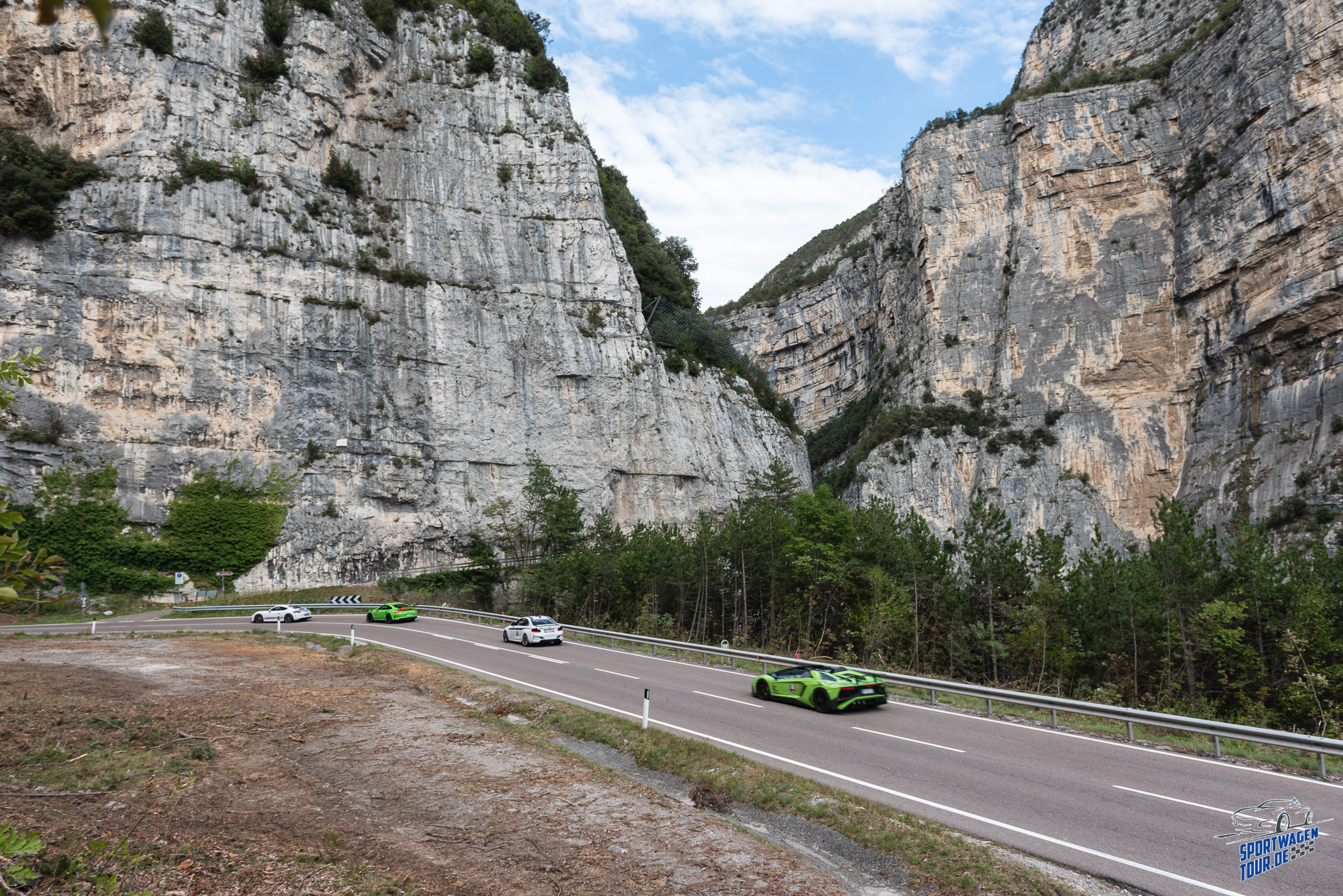 LagoRun sports cars following a road beneath towering limestone cliffs